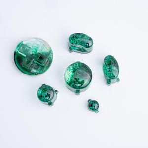 Op de afbeelding: Een set van zes doorschijnende smaragdgroene gaming-controllerknoppen. De collectie bevat een richtingspad, twee ronde knoppen, twee ovale knoppen en één kleine ronde knop, allemaal met een glanzende, gepolijste afwerking.