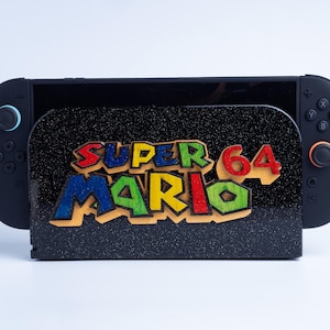 Peut inclure: Une station d'accueil pour console de jeu noire avec une finition pailletée et un logo Super Mario 64 en bois multicolore fixé à l'avant. La station est flanquée de deux manettes noires avec des accents bleus et orange sur les joysticks.