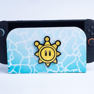 May include: A black video game console dock featuring a light blue and white water-patterned front plate with a centered yellow and black Shine Sprite emblem. The console has black controllers attached on both sides with blue and orange accents.