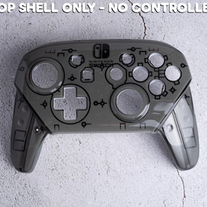 Op de afbeelding: Een grijze Nintendo Switch Pro Controller bovenste schaal. De controller heeft een transparant ontwerp met zichtbare knoppen en een D-pad. De tekst "TOP SHELL ONLY - NO CONTROLLER" staat bovenaan.