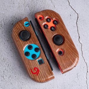 May include: Two wooden-textured game controllers with black buttons and orange and blue accents. The controllers have a wood grain pattern and feature a red logo. The buttons include directional pads and action buttons.