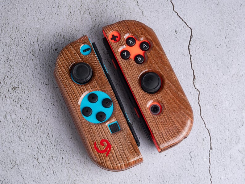 May include: Two wooden-textured game controllers with black buttons and orange and blue accents. The controllers have a wood grain pattern and feature a red logo. The buttons include directional pads and action buttons.