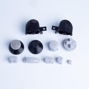 Può includere: Una collezione di parti di ricambio per controller che include due tasti dorsali neri, due coperture per levette analogiche nere e diversi pulsanti grigio chiaro etichettati con le lettere A, B, X, Y, Z e un pad direzionale su sfondo bianco.