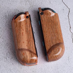 May include: Two wooden-textured Joy-Con controllers for a gaming console. The controllers have a brown wood grain pattern with a glossy finish. The design includes black, red, and blue accents. The controllers are designed to enhance the gaming experience.