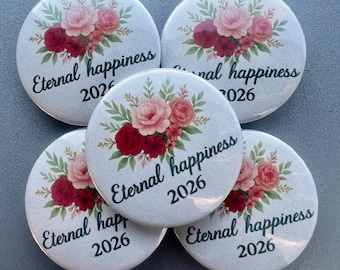 Puede incluir: Cinco botones redondos blancos con un diseño floral de rosas rosas y rojas y hojas verdes. El texto "Eternal happiness 2026" está impreso en fuente cursiva en cada botón. Los botones están dispuestos en un patrón ligeramente superpuesto.