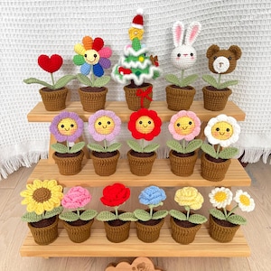 May include: A collection of crocheted flower and animal figurines in pots, displayed on a wooden shelf. The figurines include a rainbow flower, a heart, a Christmas tree, a bunny, a bear, and several smiling flowers in various colours.