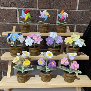 May include: A set of 10 crocheted flower arrangements in various colours, including pink, white, blue, and yellow. Each flower is potted in a small brown pot and arranged on a tiered wooden shelf.