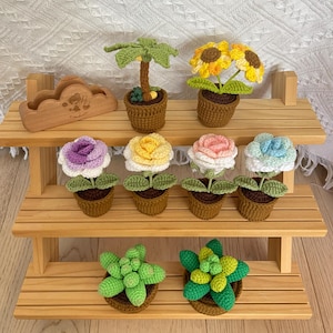 May include: A wooden tiered shelf displays a collection of handmade crochet plants. The plants include sunflowers, roses in various colours, a palm tree, and succulents, all in small brown pots. The shelf is made of light-coloured wood.