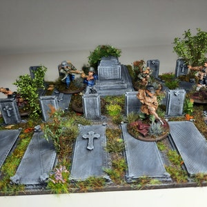 May include: A miniature wargaming terrain set depicting a graveyard with grey stone tombstones, green foliage, and miniature figures of soldiers in various poses.