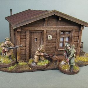 May include: A wooden cabin with a brown roof and a small window. Three miniature figures are standing outside the cabin, one holding a rifle and the other two holding guns. The cabin is on a base with green grass and brown dirt.