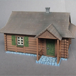 Op de afbeelding: Een miniatuur houten hutmodel met een bruin pannendak, groene luiken en een groene deur. De hut heeft een kleine veranda met een stenen fundering en een schoorsteen.
