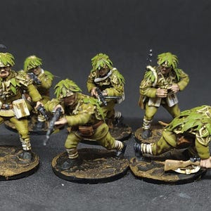 May include: Six miniature figures of soldiers wearing camouflage uniforms and carrying weapons. The figures are painted in shades of green and brown, and they are standing on small bases.