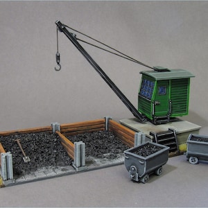 Op de afbeelding: Een groene modelkraan met een zwarte arm en een kleine cabine. De kraan staat op een platform met een stapel zwarte kolen en twee kleine kolenwagens.