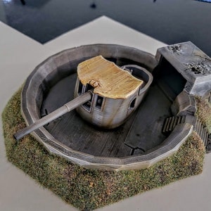 May include: A miniature model of a concrete bunker with a rotating gun turret. The turret is made of grey plastic with a gold-coloured top. The bunker is surrounded by green grass.
