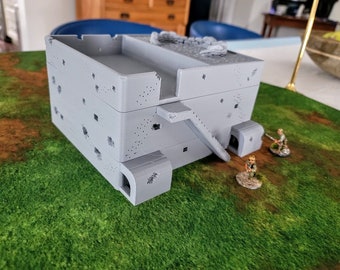 May include: A gray, multi-level, 3D printed building model with a rooftop, stairs, and multiple windows. The building is set on a green grassy surface with two small, gray figures standing in front of it.