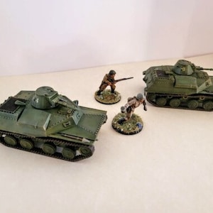 Puede incluir: Dos tanques militares verdes con detalles pintados y dos figuras en miniatura de soldados con uniformes de camuflaje. Las figuras sostienen rifles y están de pie sobre pequeñas bases.