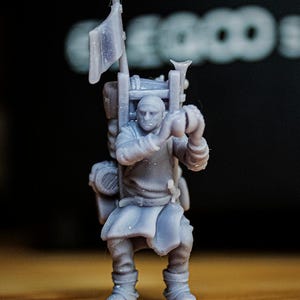 Op de afbeelding: Een kleine, lichtgrijze figuur van een persoon met een rugzak en een vlag. De figuur is een fantasiekarakter. De figuur staat op een houten oppervlak. De achtergrond is wazig.