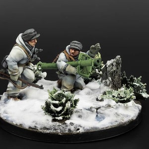 Könnte beinhalten: Zwei bemalte Miniaturfiguren von Soldaten in Wintertarnkleidung, einer mit einem Gewehr und der andere mit einem Raketenwerfer, sind in einer Schneelandschaft mit kleinen immergrünen Bäumen in Szene gesetzt.