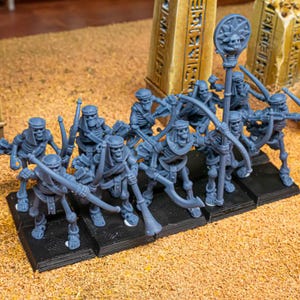 Op de afbeelding: Een groep grijze miniatuur skeletboogschutters met pijl en boog, staand op zwarte bases. Eén figuur houdt een staf vast met een schedel en zonnester ontwerp. De figuren staan tegen een achtergrond van beige zand en oude pilaren.