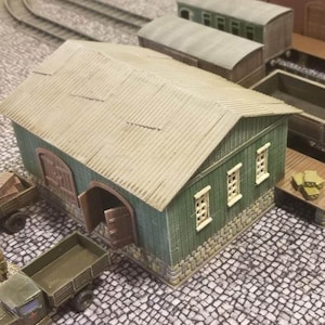 May include: Miniature model of a green and tan building with an open door, part of a model train set. The building has a corrugated roof and is surrounded by other train cars and trucks. The scene is set on a cobblestone surface.