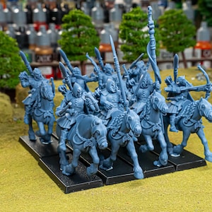 Puede incluir: Un grupo de figuras en miniatura azules de soldados montados, cada uno con una lanza o arco, dispuestos sobre una base negra. Los caballos tienen crines y bridas detalladas. El fondo incluye árboles en miniatura y botes de pintura.