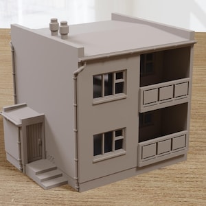 May include: A miniature model of a two-story building in a neutral beige color. The building features windows, balconies, a small entrance, and a flat roof with two small chimneys. The model is set on a wooden surface.