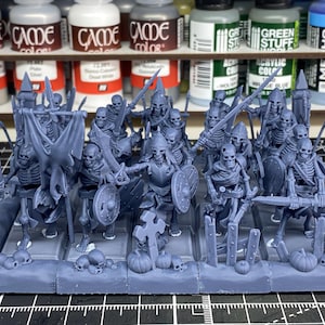 Puede incluir: Un grupo de miniaturas grises de guerreros esqueleto, armados con espadas, escudos y lanzas. Algunas figuras llevan una bandera. Las miniaturas están sobre una base rectangular con calaveras y calabazas. Hay botes de pintura en el fondo.