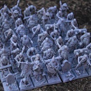 Puede incluir: Colección de figuras en miniatura grises, probablemente para juegos de mesa. Las figuras representan varios personajes de fantasía, incluyendo zombis y guerreros, dispuestos sobre bases individuales. La escena se desarrolla sobre una superficie texturizada.