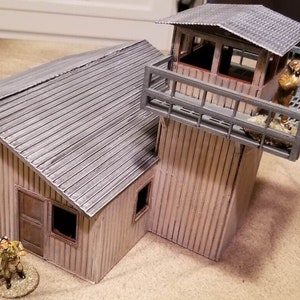 May include: A model of a building with a watchtower. The building is made of wood and has a corrugated metal roof. The watchtower has a railing and a figure standing on it. The building is painted in a light brown colour and the watchtower is painted in a dark grey colour.