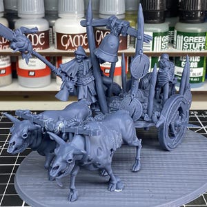 May include: An unpainted miniature model of a fantasy war chariot, pulled by two horned creatures. The chariot carries figures, a bell, and shields. The model is on a round base, with paint bottles in the background.