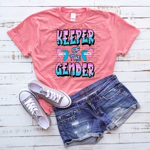 Może przedstawiać: R&oacute;żowa koszulka z napisem "Keeper of the Gender" w stylu graffiti. Tekst jest otoczony dwoma niebieskimi i r&oacute;żowymi śladami st&oacute;p.