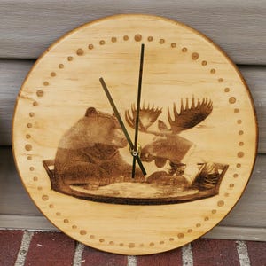 Puede incluir: Un reloj de madera redondo con acabado marrón claro. La esfera del reloj presenta una imagen detallada grabada en madera de un oso y un alce. El reloj tiene manecillas negras y pequeñas marcas punteadas alrededor del borde.