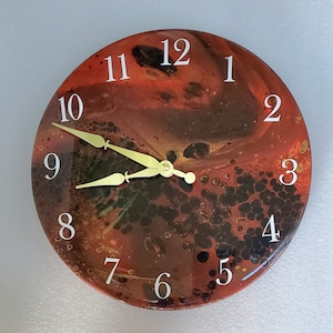 May include: Round wall clock with a red, brown, and black abstract design. The clock face features white numerals and gold-colored hands. A unique decorative piece for any room.