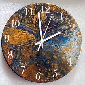 May include: Round wall clock with a unique abstract design in shades of gold, blue, and brown. The clock face features white numerals and white clock hands. The clock's design has a marbled effect.
