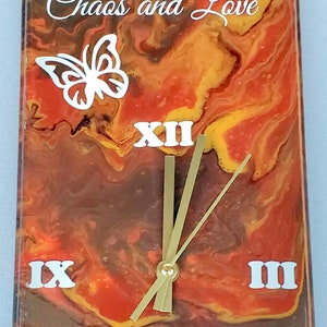 Puede incluir: Reloj de pared rectangular con un fondo arremolinado naranja, marrón y amarillo. La esfera del reloj presenta números romanos blancos y el texto "Our Family Is Just The Right Mix Of Chaos and Love". También hay un gráfico de mariposa blanca.