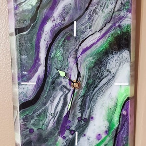 Puede incluir: Un reloj con un diseño abstracto verde, morado y negro. La esfera del reloj es blanca con agujas negras.