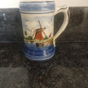 May include: A blue and white ceramic beer stein featuring a painted scene of a windmill in a Dutch landscape. The handle is white with a blue rim.