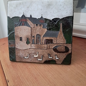 May include: A square ceramic tile featuring a textured relief design of a castle with dark green hills in the background. The foreground depicts a light brown courtyard with five white geese swimming in a small pond. The sky is a mottled white.
