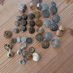 May include: A collection of assorted metal buttons in various shapes, sizes, and finishes, including silver, gold, and bronze tones. The buttons feature intricate embossed patterns, geometric textures, and smooth surfaces, with some displaying circular designs.