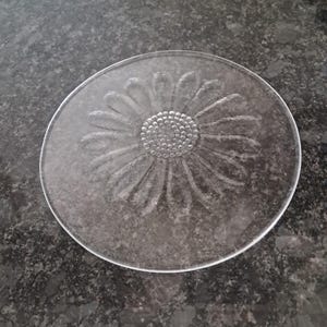 May include: A clear glass coaster featuring a daisy design etched into the surface. The coaster is circular with a raised edge. The daisy has detailed petals and a textured centre.