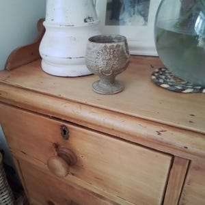 Könnte beinhalten: Ein kleines, verziertes, beigefarbenes Keramikkelch mit detailliertem Blumendesign steht auf einer Holzkommode. Ein weißer Krug und eine Glasschale mit Wasser und Pflanzen befinden sich ebenfalls auf der Kommode. Die Kommode hat zwei Schubladen und einen Holzknauf.