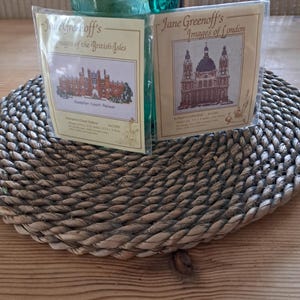 May include: Two cross-stitch kits showcasing British landmarks. One shows Hampton Court Palace, the other St. Paul's Cathedral. The kits are in clear packaging, resting on a woven mat. A green glass bottle is in the background.