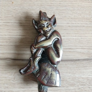 Könnte beinhalten: Ein Türklopfer aus Messing in Form eines grinsenden Kobolds, der auf einem Felsen sitzt. Der Kobold trägt einen spitzen Hut und hält seine Beine fest.