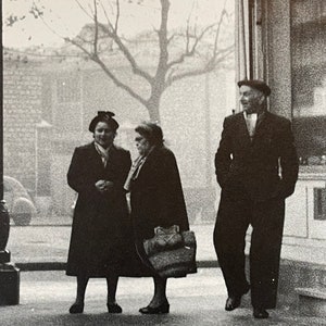 May include: Three people, two women and one man, standing on a pavement in front of a building. The women are wearing hats and coats. The man is wearing a hat and a suit. The image is in black and white.
