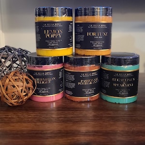 May include: Five glass jars of body sugar scrub with black labels. The jars are arranged in two rows. The labels have different colors and feature the text "La Bella Body" and the names of the scents: Lemon Poppy, Fortune, Watermelon Delight, Passionfruit & Pomegranate, Eucalyptus & Spearmint.