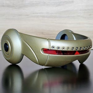Puede incluir: Un dispositivo dorado de aspecto futurista, con un extremo circular y un cuerpo curvado. Presenta un acento rectangular rojo con pequeños detalles circulares y una lente negra. El objeto está sobre un fondo de madera.