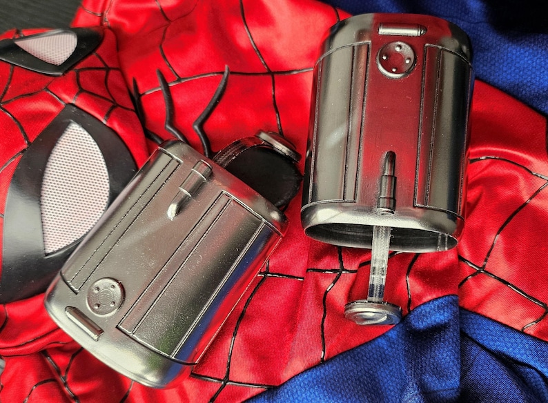 May include: Two silver wrist-mounted devices with detailed paneling and a clear, cylindrical component extending from one. The devices rest on a red and blue fabric with a spiderweb pattern, suggesting a superhero costume.