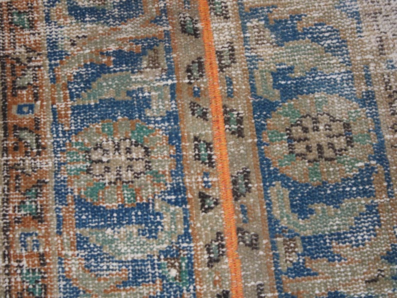 May include: Close-up of a blue and brown patterned rug with a detailed design. The rug has a fringe border and a subtle orange stripe running down the center.