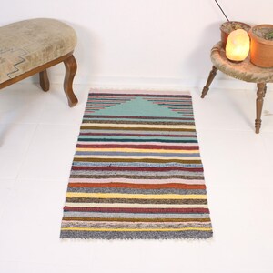 Puede incluir: Una alfombra rectangular tejida a mano con un patrón de rayas en varios colores, incluyendo turquesa, rojo, amarillo y gris. La alfombra tiene un diseño triangular en la parte superior y un borde con flecos. La alfombra mide aproximadamente 91 cm de largo y 61 cm de ancho.