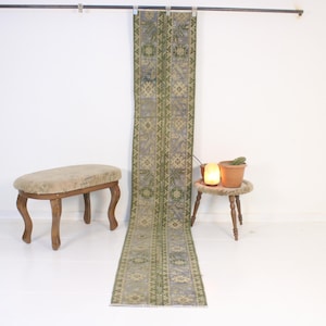 Peut inclure: Un long tapis rectangulaire avec un motif géométrique répétitif dans les tons de vert, de bleu et de beige. Le tapis est exposé verticalement contre un mur blanc. Une petite table ronde en bois avec une plante en pot et une lampe est à droite.